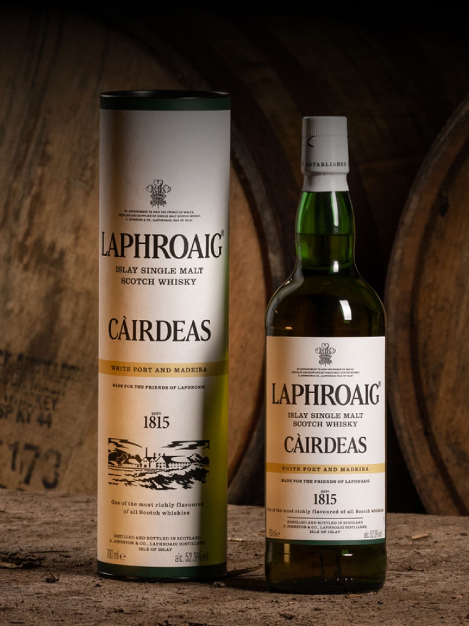 Bottle of Laphroaig Càirdeas 2023 White Port & Madeira with the gifting sleeve it comes in sitting in front of barrels