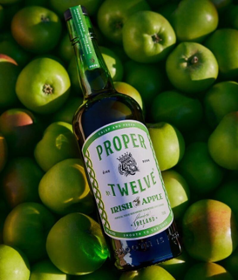 A bottle of Proper No. Twelve™️ Irish Apple laying on top of green apples