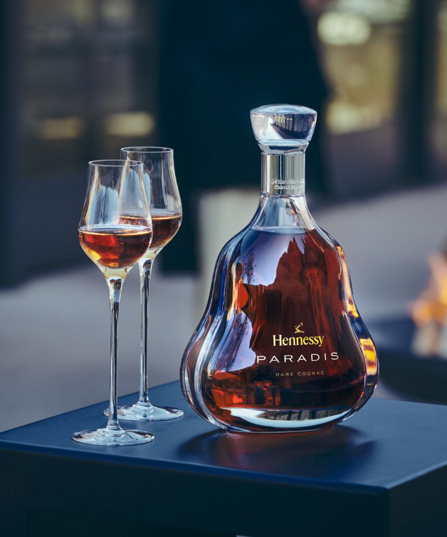 Hennessy Paradis Rare Cognac outside by a fire pit, two toasting flutes beside it.