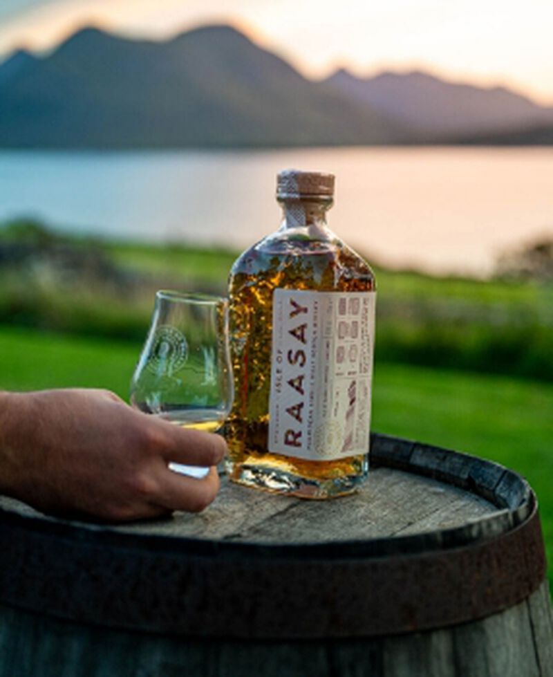 A bottle of Isle of Raasay Hebridean Single Malt Scotch with a glass sitting on a barrel outside with water and mountains in the background