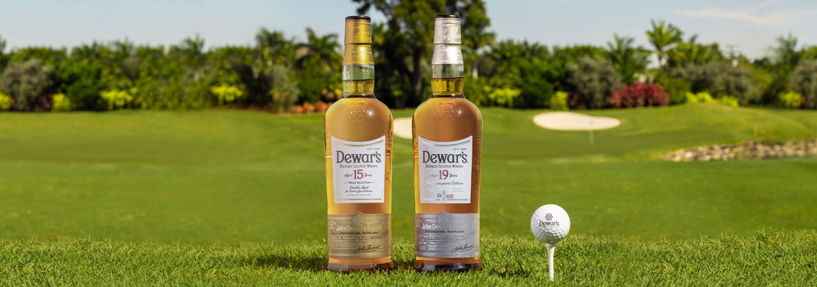 Dewar's Aged 15 Years and Aged 19 Years bottles beside a golf ball on a tee on golf course turf