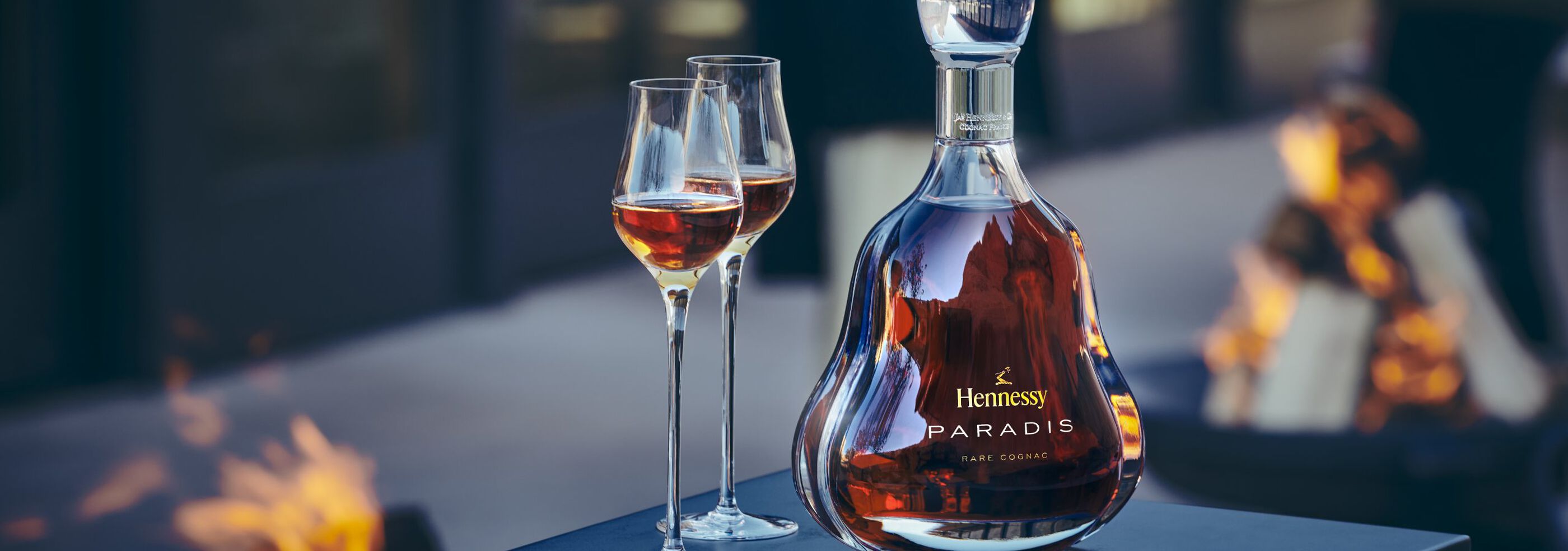 Hennessy Paradis Rare Cognac outside by a fire pit, two toasting flutes beside it.