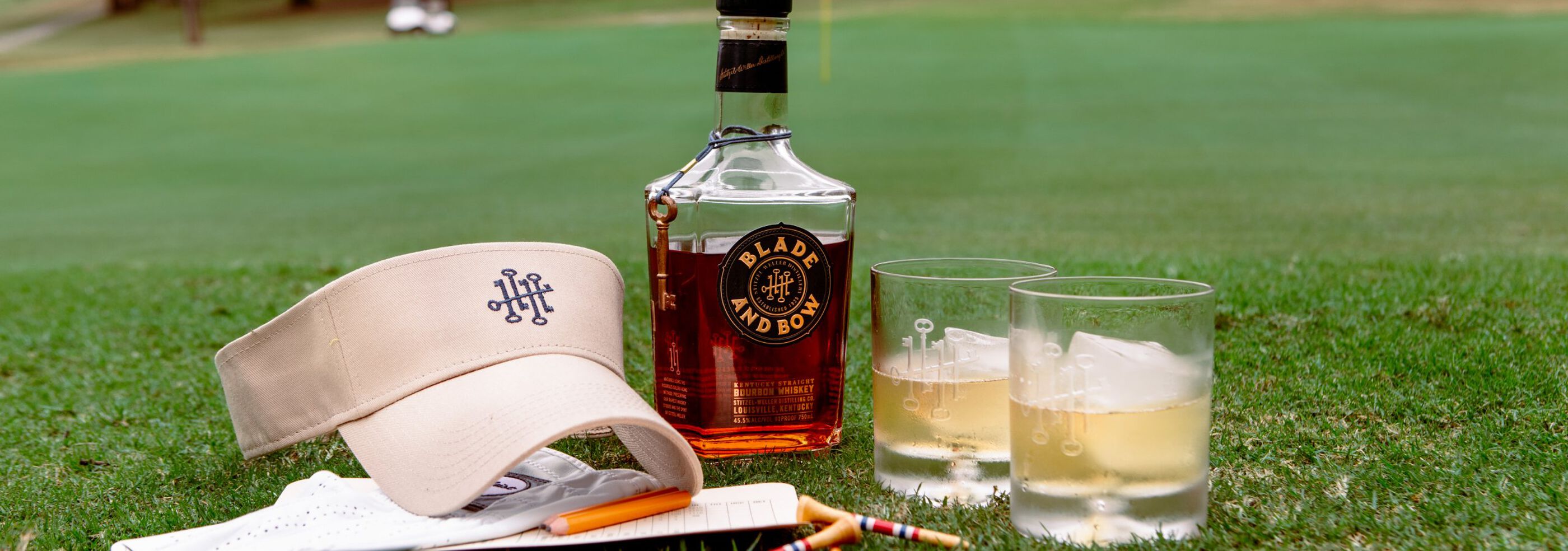 Blade and Bow bottle on golf course turf, beside it- two rocks glasses, golf tees, a visor, gloves, and scorecard