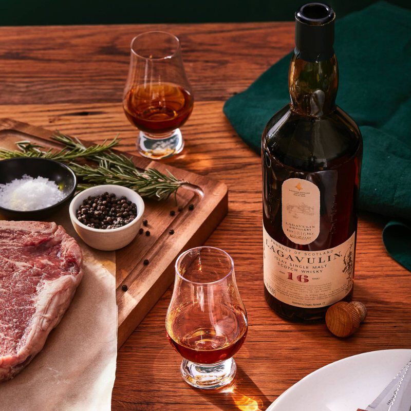 Lagavulin 16 Year Old Islay Single Malt Scotch Whisky beside a cutting board with steak, salt, rosemary, peppercorns. Beside it, two glencairn style glasses with scotch.